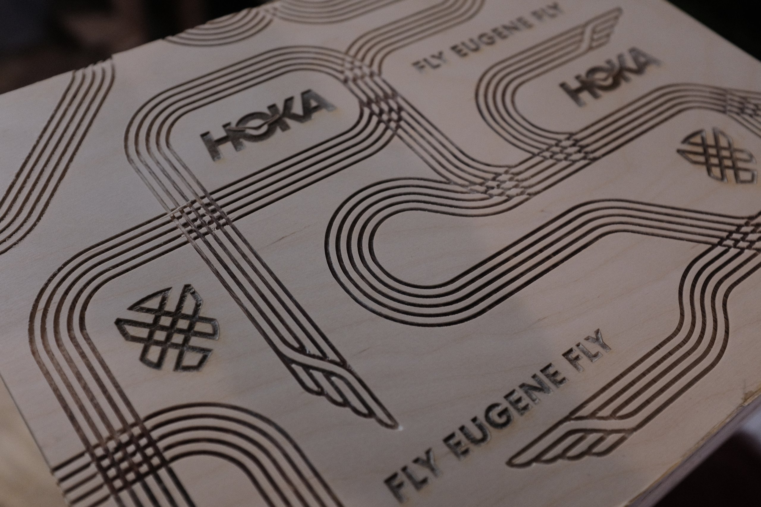 Laser-engraved plywood panel with HOKA logos and "FLY EUGENE FLY" track-line patterns