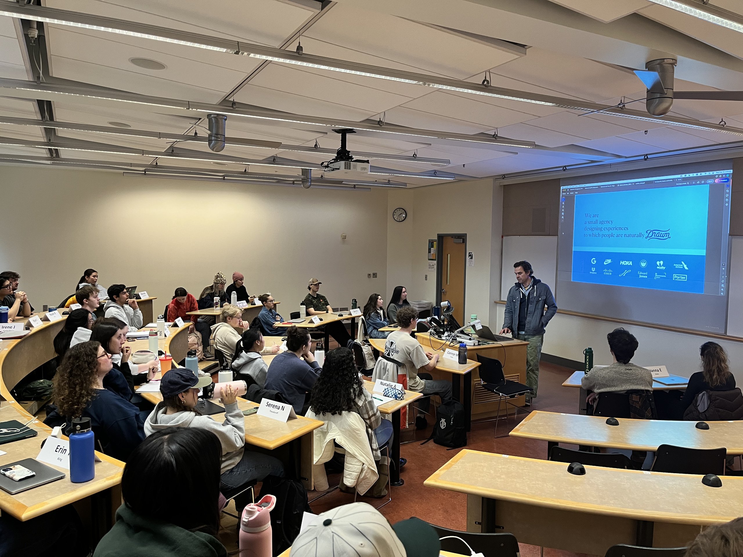 Drawn presenter addressing a university classroom; slide shows agency tagline and client logos