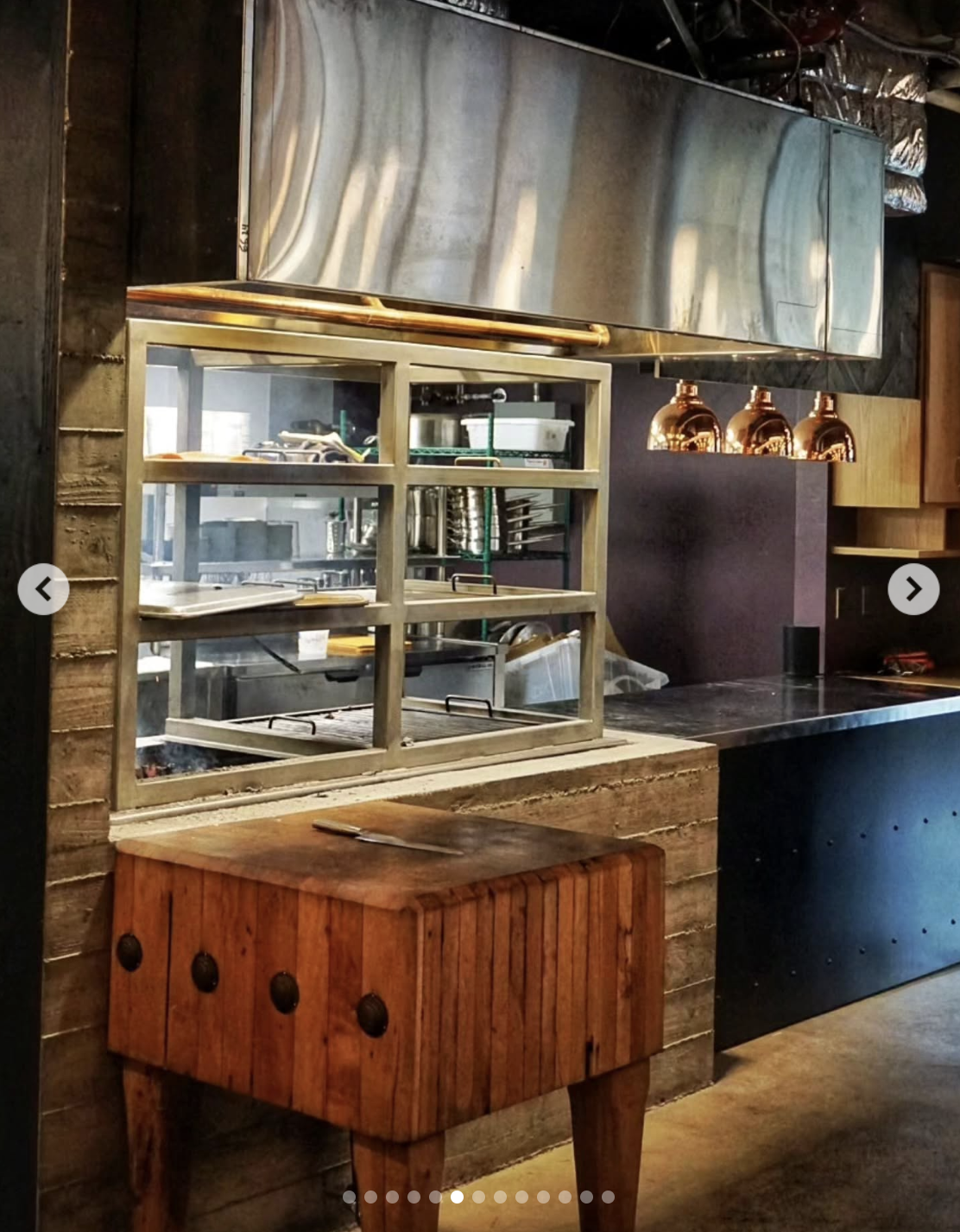 Dark restaurant kitchen with glass-front charcoal grill, copper heat lamps, and wooden butcher block