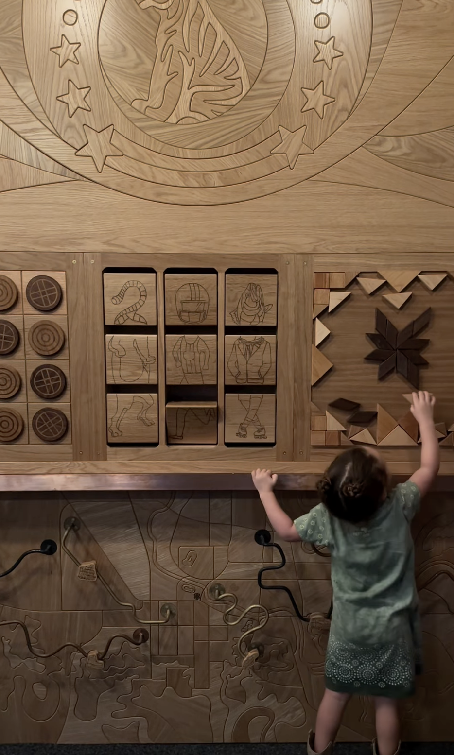 Child reaching up to a carved wood interactive wall with engraved animals and shapes