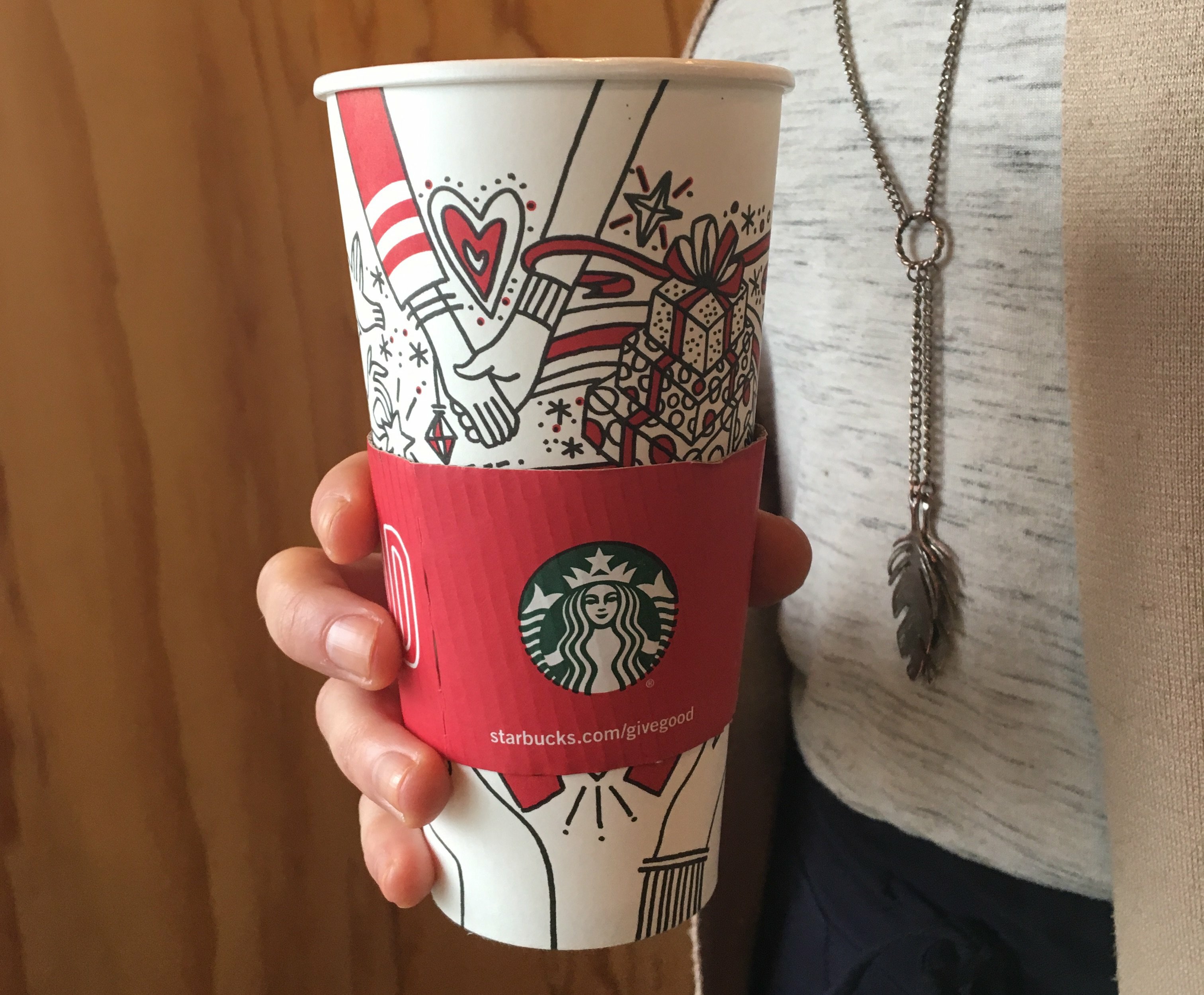 Hand holding an illustrated Starbucks holiday cup in a red Give Good sleeve with the siren logo