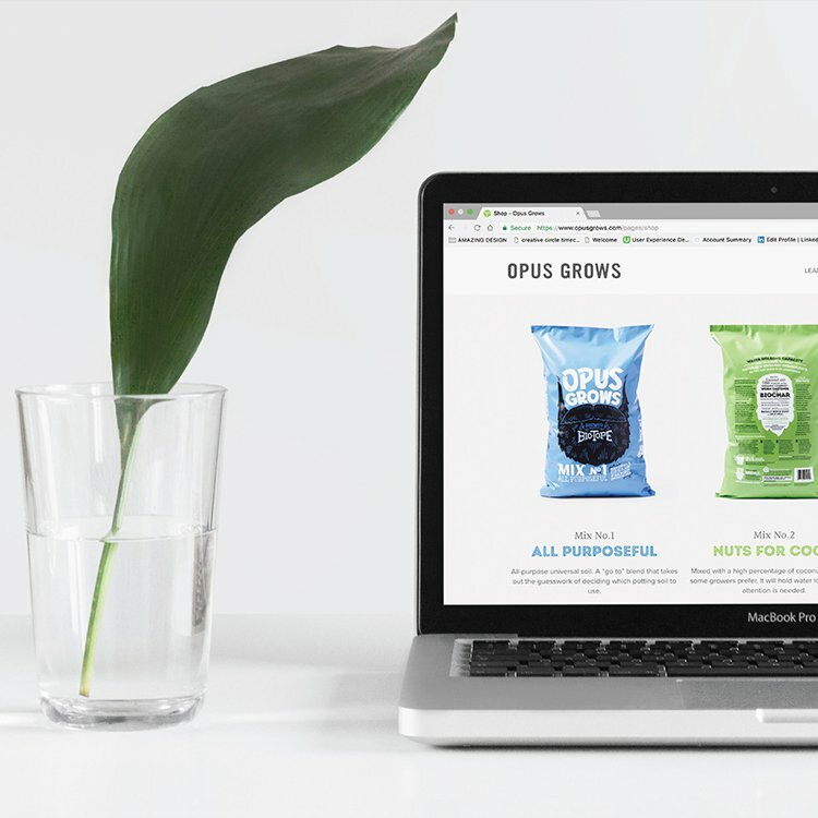 Laptop showing the Opus Grows website with soil product bags beside a leaf in a water glass