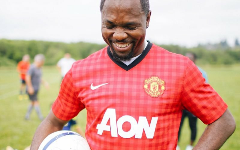 Smiling man in a red Manchester United AON jersey holding a soccer ball on a field, by Trask Bedortha