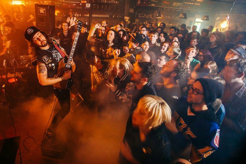 Guitarist performing for a packed, smoky crowd at a dim bar, by Trask Bedortha