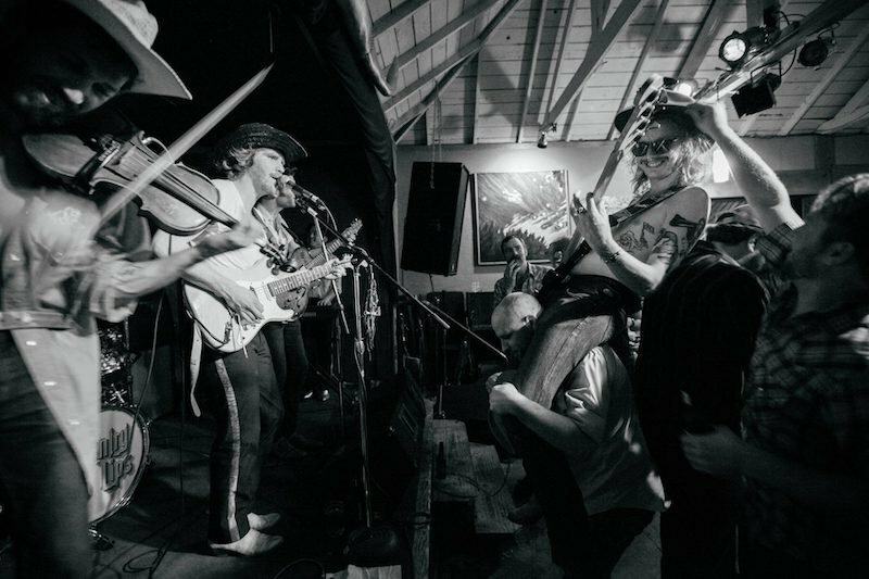 Black-and-white shot of a band with fiddle and guitar playing to a dancing crowd, by Trask Bedortha