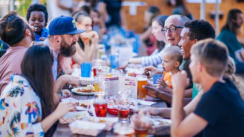 Families with children sharing a meal and drinks at a long communal table outdoors