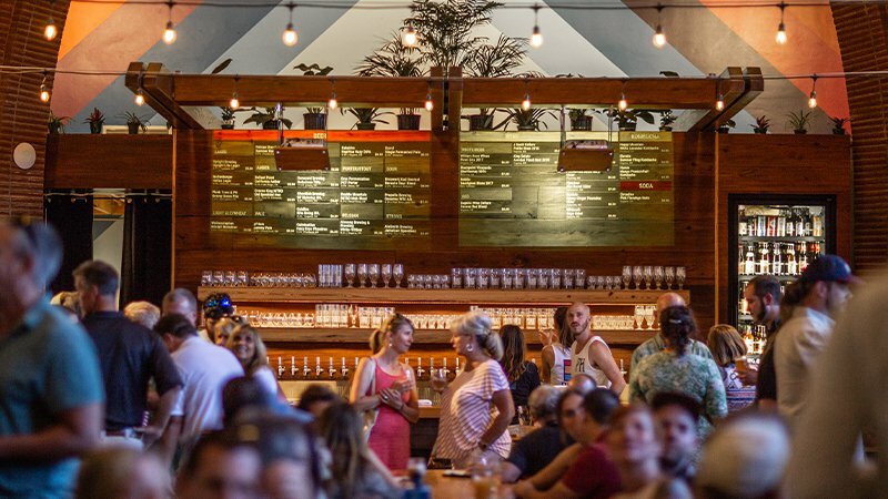 Busy restaurant interior with patrons at communal tables below glowing menu boards