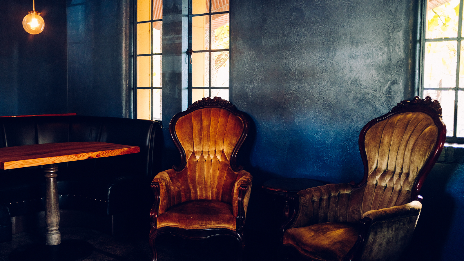 Two antique velvet upholstered armchairs and a wood table by tall windows in a dim moody whiskey lab