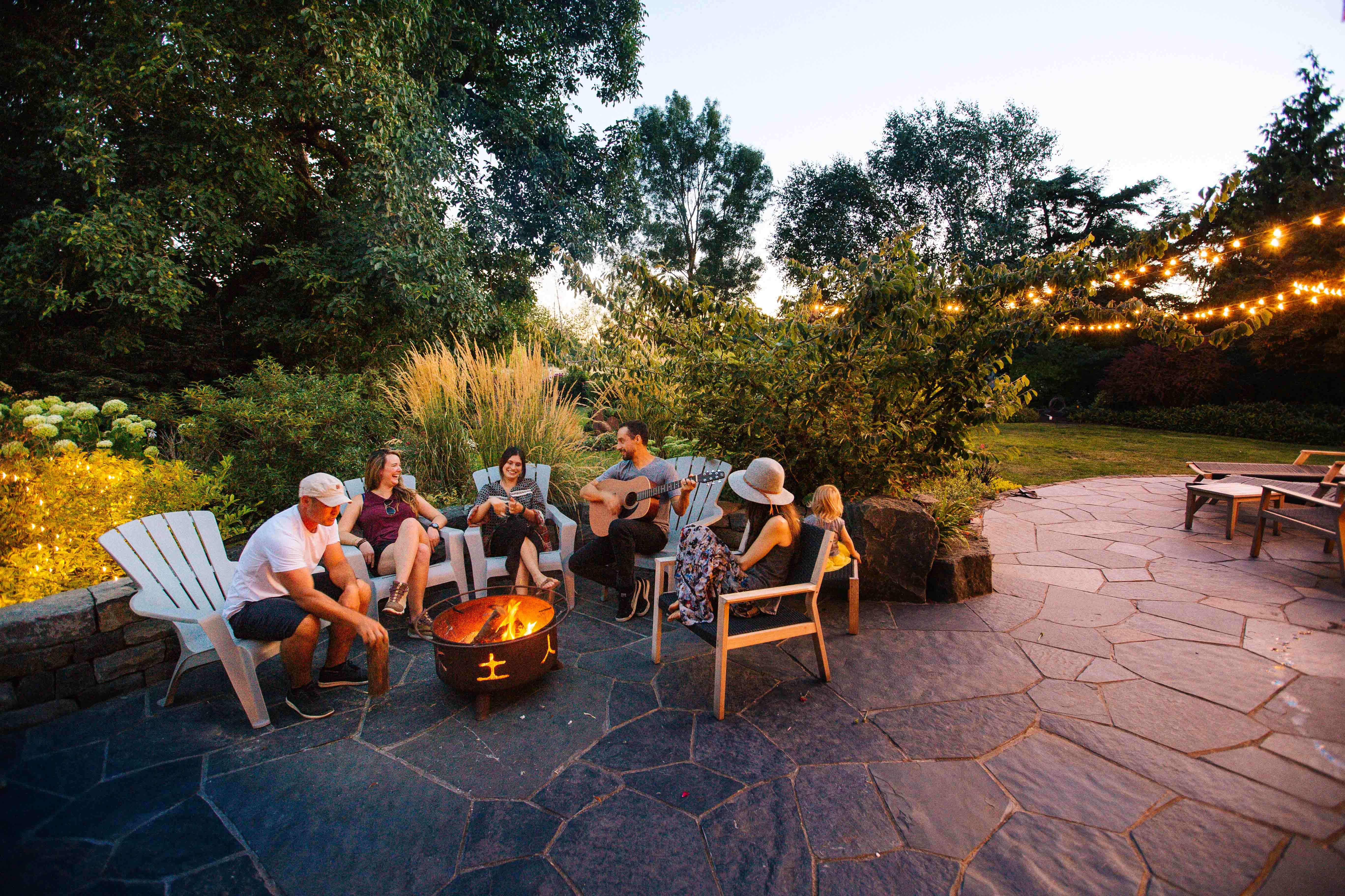 Group relaxing in chairs around a backyard fire pit under string lights at dusk