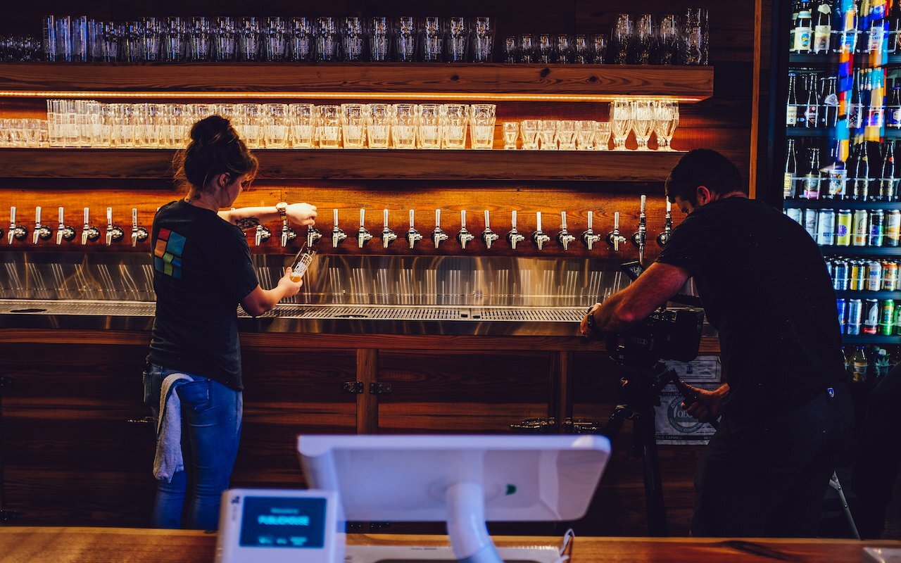 Two staff working behind a wood bar lined with taps and shelves of glassware and bottles