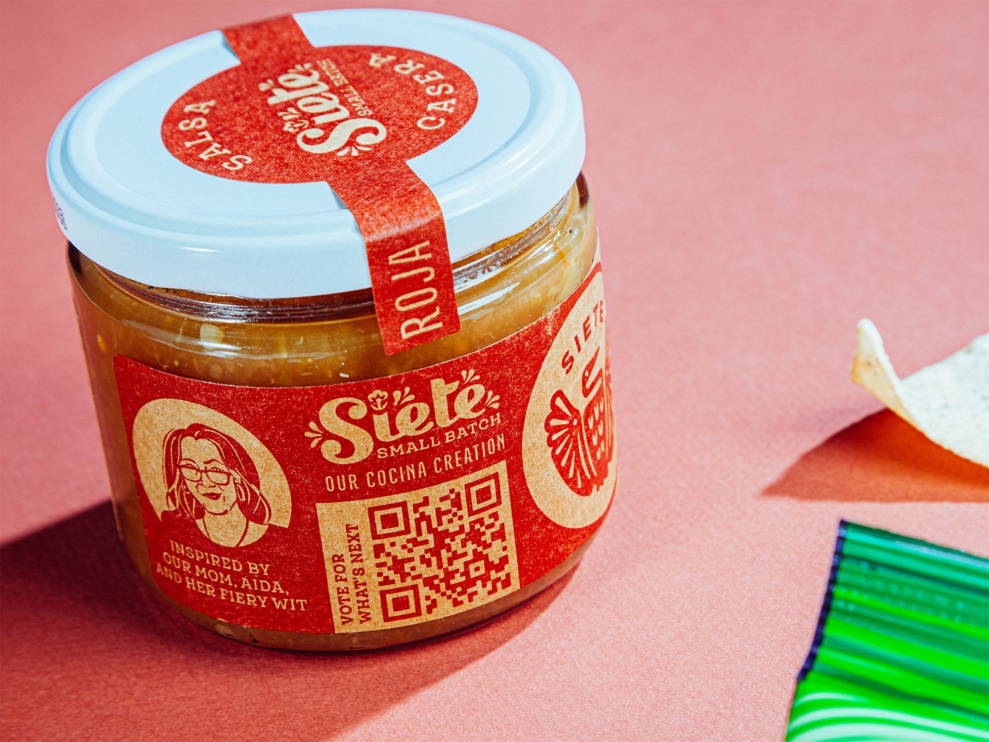 Siete Small Batch Salsa Roja jar with QR code label and tortilla chip on a pink background