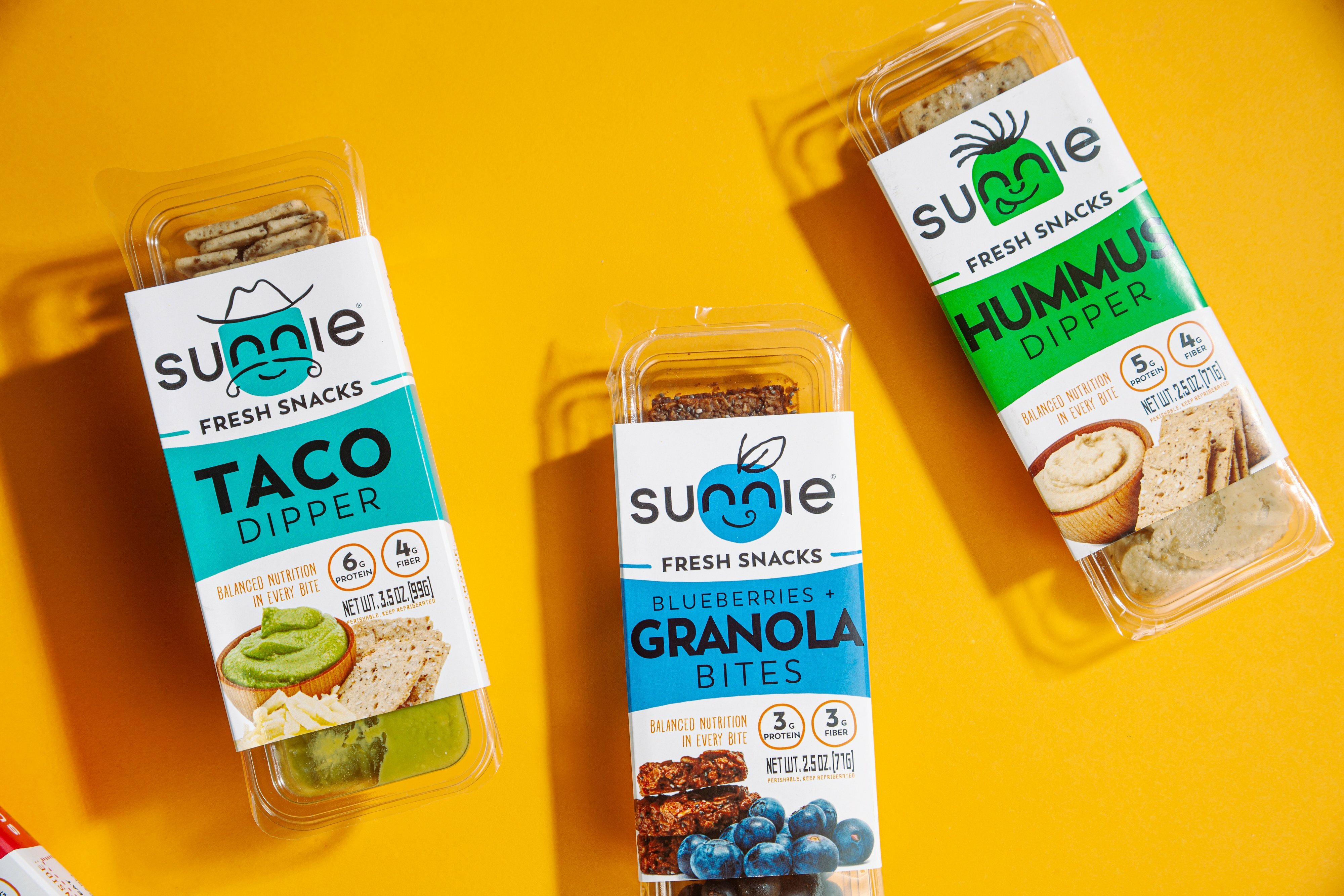 Three Sunnie Fresh Snacks packs - Taco Dipper, Blueberries + Granola Bites, and Hummus Dipper - on yellow