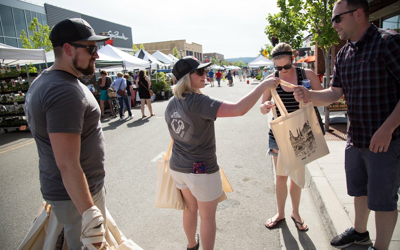 People exchanging a printed tote bag at an outdoor street market on a sunny day