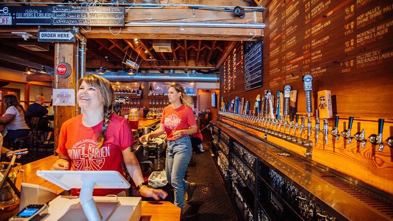 Bartenders in red Growl + Garden shirts at a wooden bar lined with beer taps