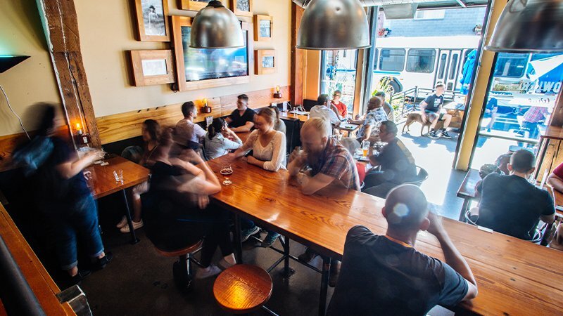 Patrons drinking and talking at long wooden tables inside a busy taproom