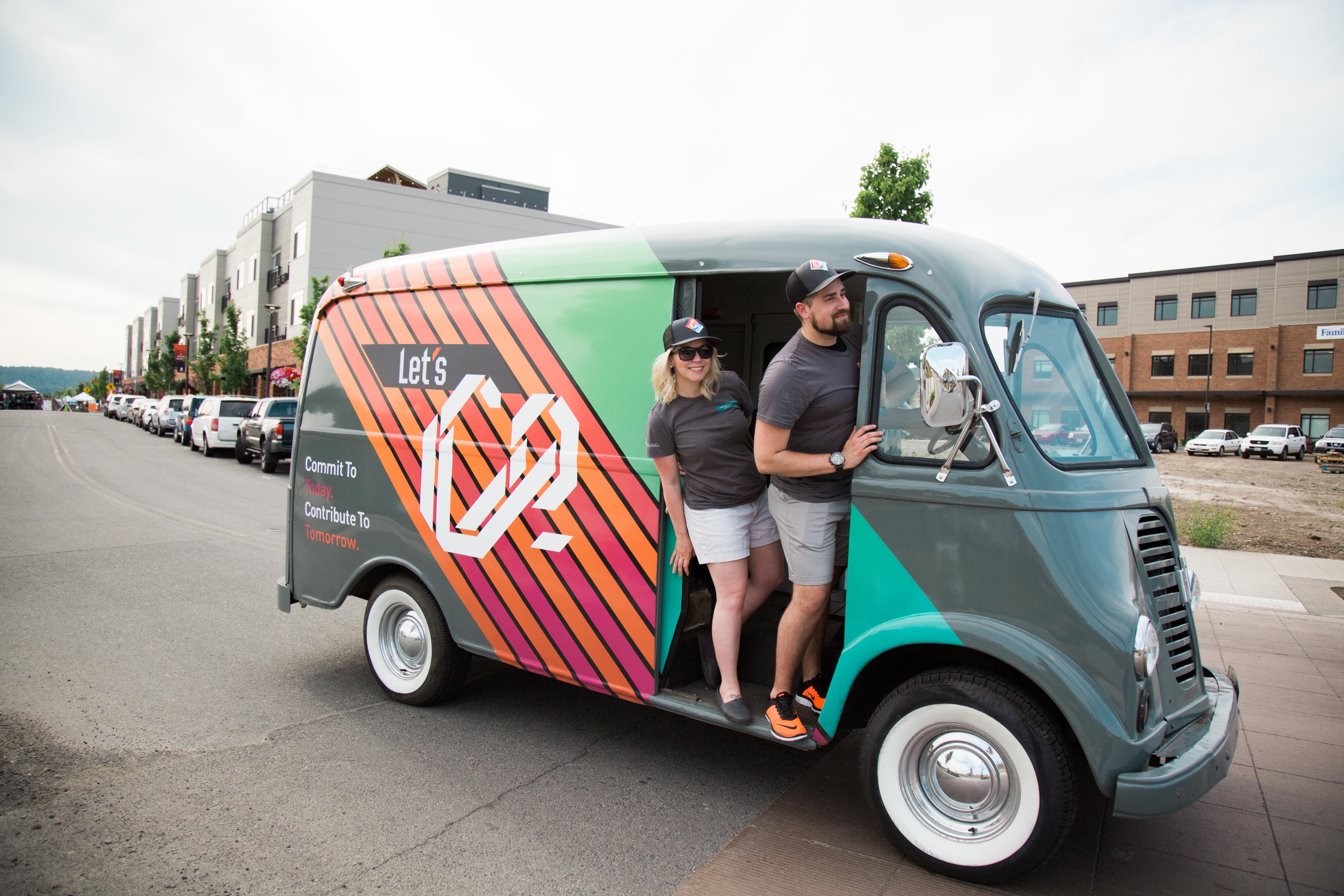 Two people standing in a vintage delivery van wrapped with WSECU Let's Contribute heart graphics